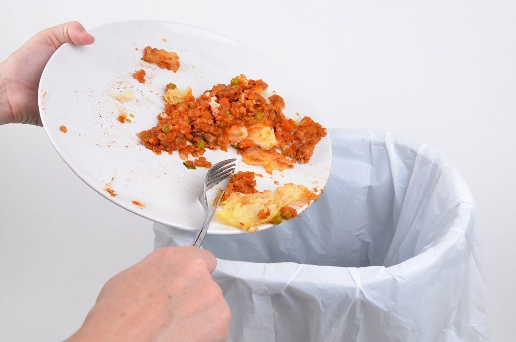 Person scraping food waste from a dinner plate into a trash bin.